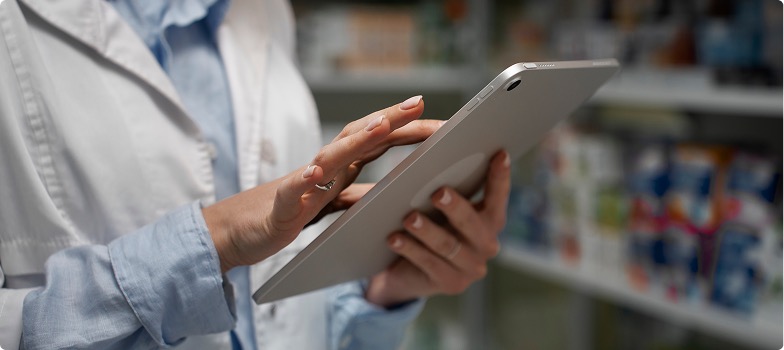 Pharmacy professional using a tablet in front of shelves.